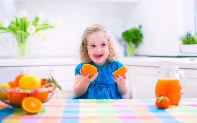 A young girl playfully eats oranges at a kitchen table
