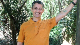 Luis Souza smiles while resting an arm against a tree