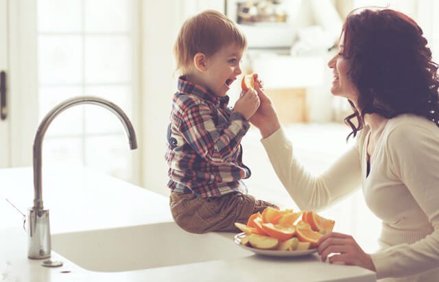 A mother feeds her young boy oranges by a kitchen sink