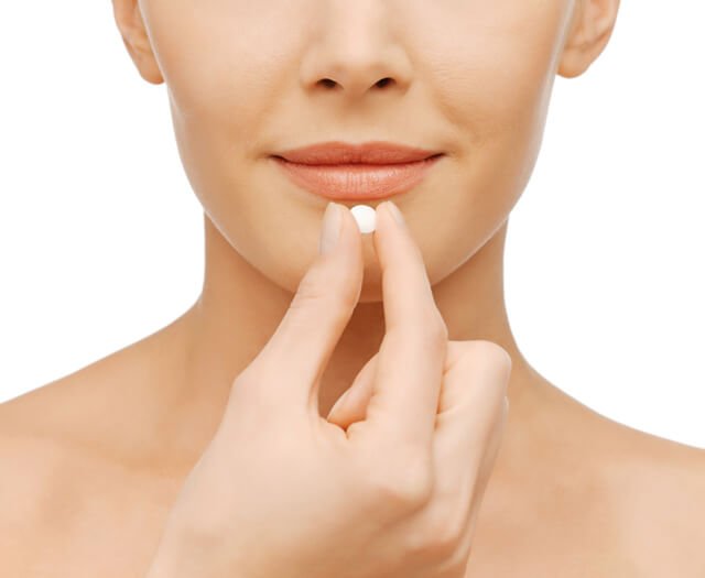 A woman holds a pill in front of her mouth against a white background