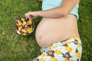 A pregnant woman eats fruit from a bowl on a lawn