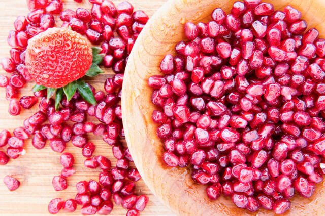 Wooden bowl containing pomegranate seeds and a single strawberry