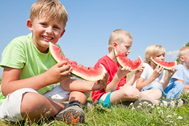 Boys eat large watermelon slices on grass under a bright blue sky