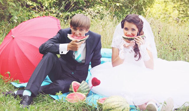 A bride and groom eat large watermelon slices while sitting on a blanket on grass